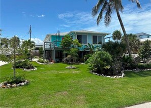 A two-story house with a green lawn, palm trees, and a small garden with rocks and plants. - The Blue Retreat in Fort Myers Beach (Fort Myers Beach)