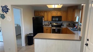 A compact kitchen with wooden cabinets, a microwave, a stove, and a trash bin. - The Blue Retreat in Fort Myers Beach (Fort Myers Beach)