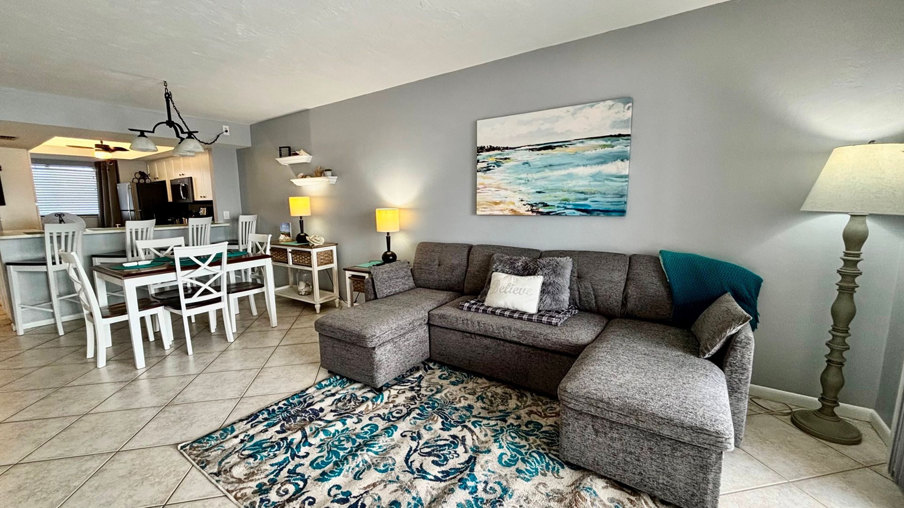 A living room with a grey sectional sofa, a dining table with chairs, a patterned rug, and a painting of a seascape.