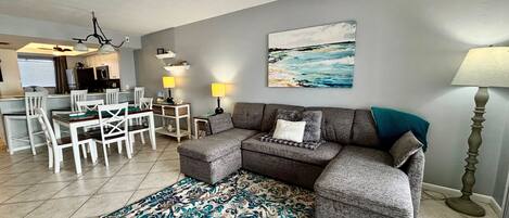 A living room with a grey sectional sofa, a dining table with chairs, a patterned rug, and a painting of a seascape.