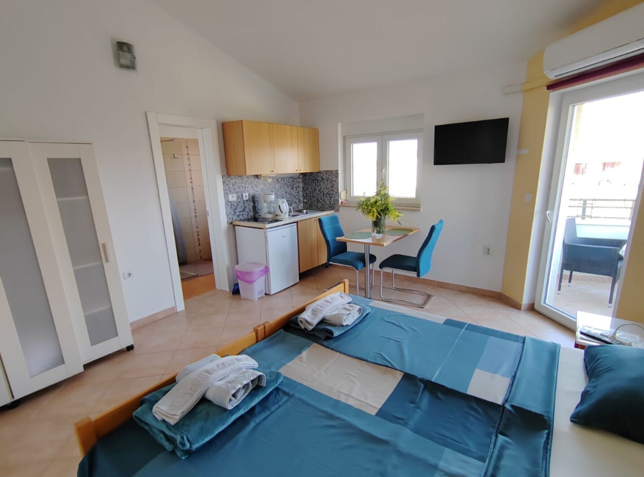 A compact room with a kitchenette, a bed with blue bedding, a small table with chairs, and a TV mounted on the wall.