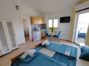 A compact room with a kitchenette, a bed with blue bedding, a small table with chairs, and a TV mounted on the wall. - Apartments Lighthouse -(A4) - TwoBedroom Apartment with Sea View (Fazana)