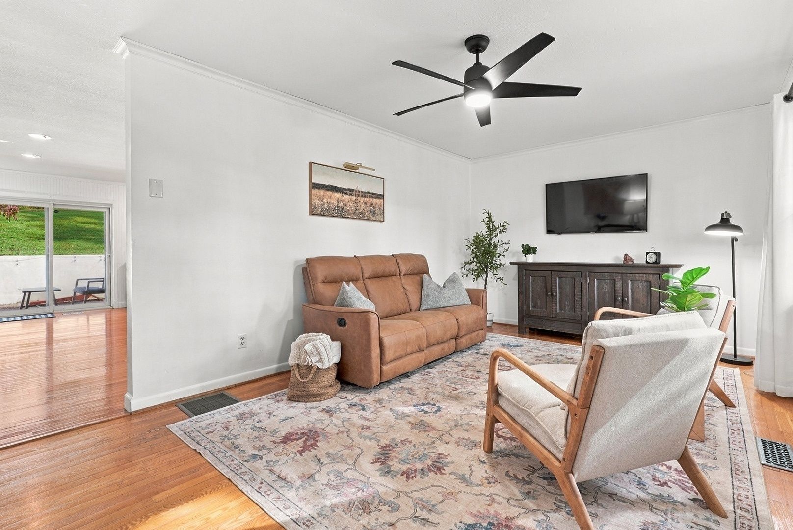 A living room with a brown sofa, a wooden chair, a patterned rug, a ceiling fan, and a TV on the wall.