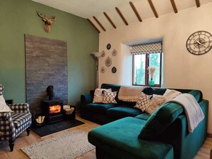 Un salón acogedor con chimenea, un sofá verde, un sillón estampado y decoración rústica en la pared.