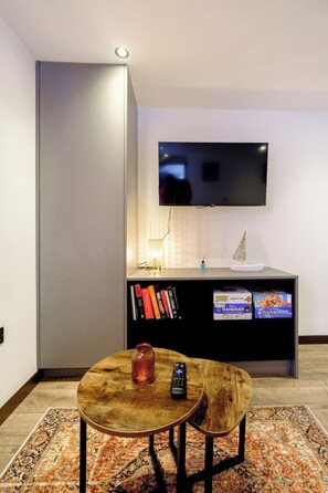 Ein modernes Wohnzimmer mit einem an der Wand befestigten Flachbildfernseher, einem Holztisch mit Fernbedienung und einer Glasvase sowie einem Bücherregal mit verschiedenen Gegenständen, darunter Bücher und Gesellschaftsspiele.