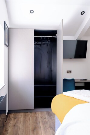 Ein modernes Hotelzimmer mit begehbarem Kleiderschrank, einem Bett, einem Schreibtisch mit Stuhl und einem flachbildfernseher an der Wand.