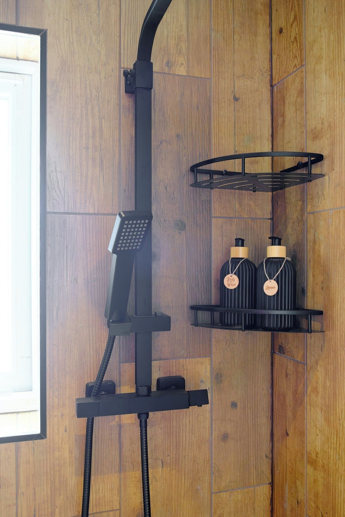 A shower area with a handheld showerhead, a black towel rack, and two black bottles with labels on a wooden wall.