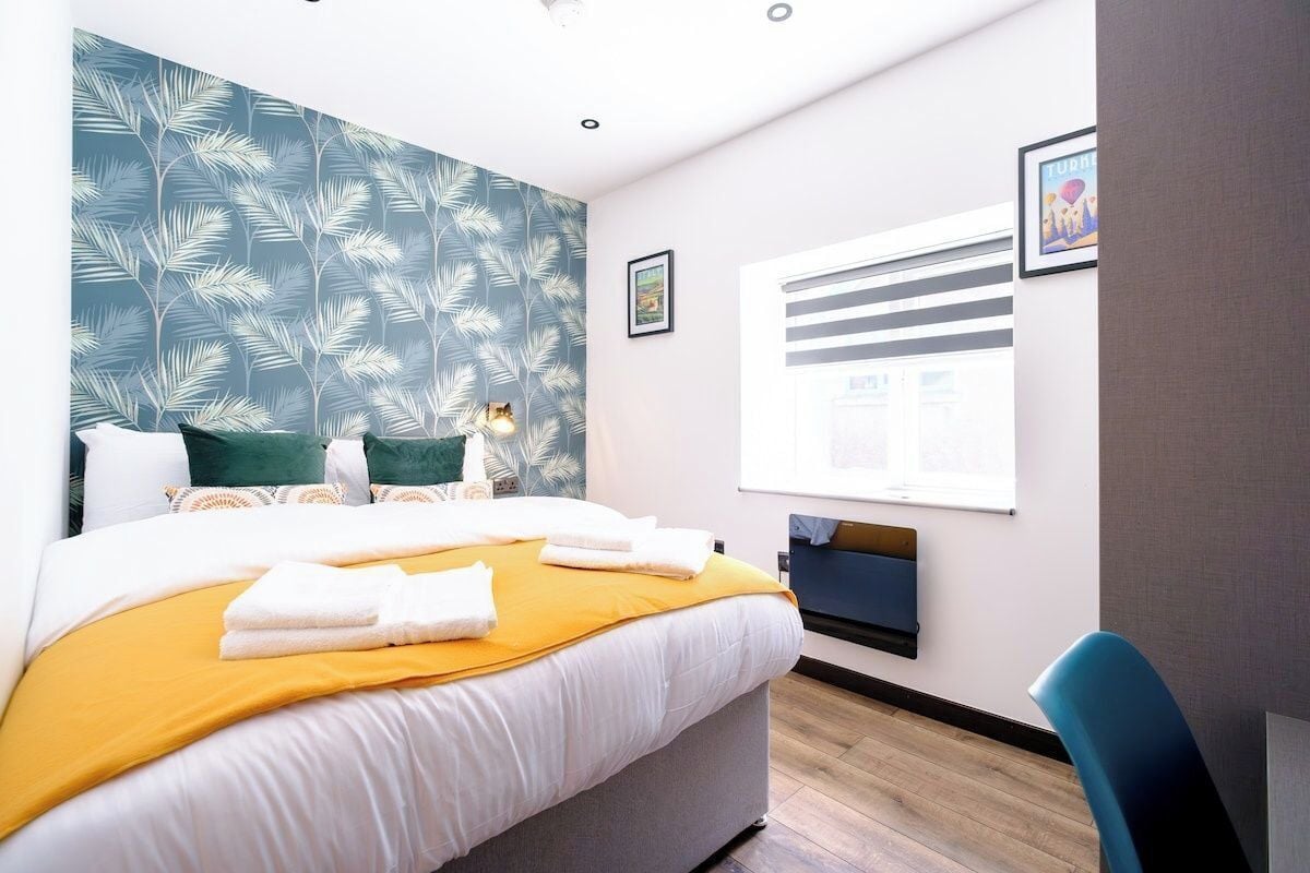 A hotel room with a bed, a desk, a chair, a window, and a wall with a leaf pattern.