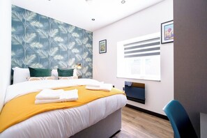 Ein Hotelzimmer mit einem Bett, einem Schreibtisch, einem Stuhl, einem Fenster und einer Wand mit einem Blattmuster.