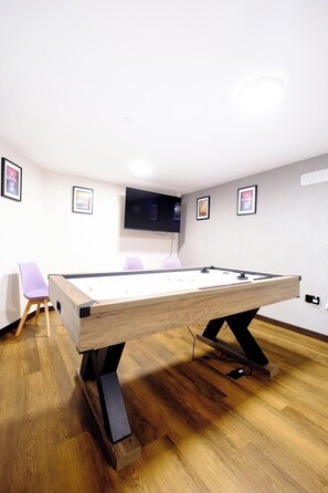 Une table de billard dans une pièce où l’on trouve un téléviseur à écran plat fixé au mur, un fauteuil violet et des affiches encadrées accrochées aux murs.