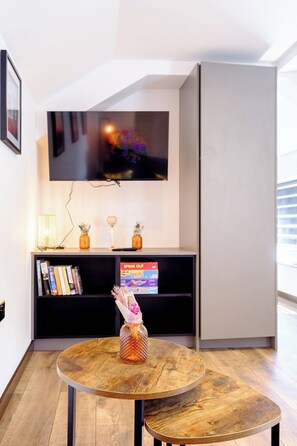 Een moderne woonkamer met een flatscreen televisie aan de muur, een boekenkast met boeken en decoratieve objecten, en een ronde houten tafel met een vaas.