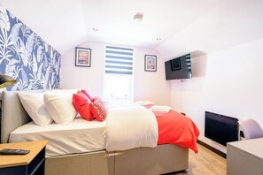 Une chambre avec un lit, une télévision, une fenêtre avec des stores et un mur orné d’un motif tropical.