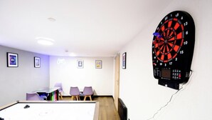 Eine an der Wand befestigte Dartscheibe, ein Billardtisch und eine Sitzecke mit Stühlen.
