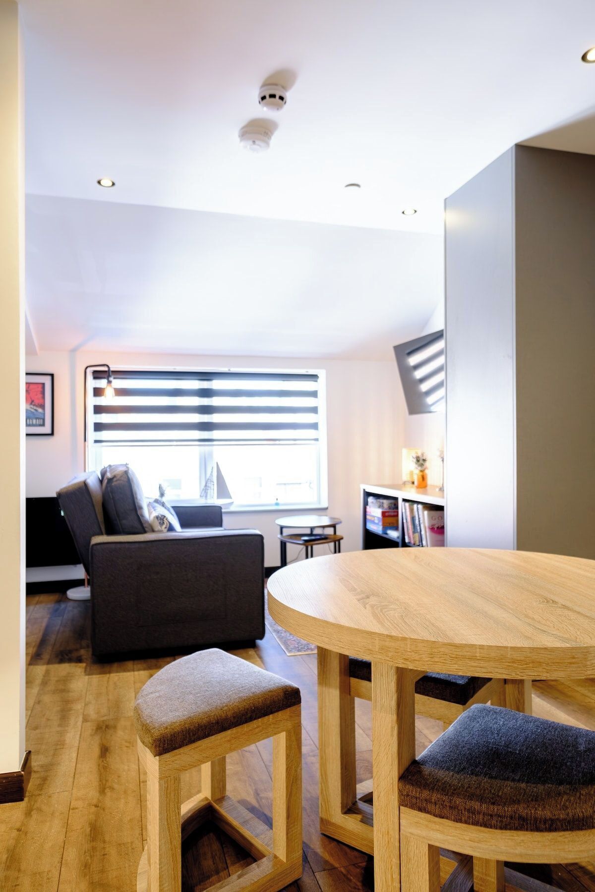 Un salon moderne avec une table ronde en bois, deux tabourets assortis, un canapé foncé et une fenêtre avec des stores.