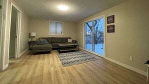 Ein Wohnzimmer mit einer Eckcouch, einem kleinen Tisch, einer Lampe, einem gemusterten Teppich und einer Glastür, die zu einer verschneiten Außenanlage führt.