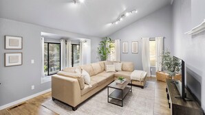 A spacious living room with a beige sectional sofa, a coffee table, and a flat-screen TV. There are large windows providing natural light and a view of greenery outside. - Two Living Spaces • Private Downstairs Suite (Grants Pass)