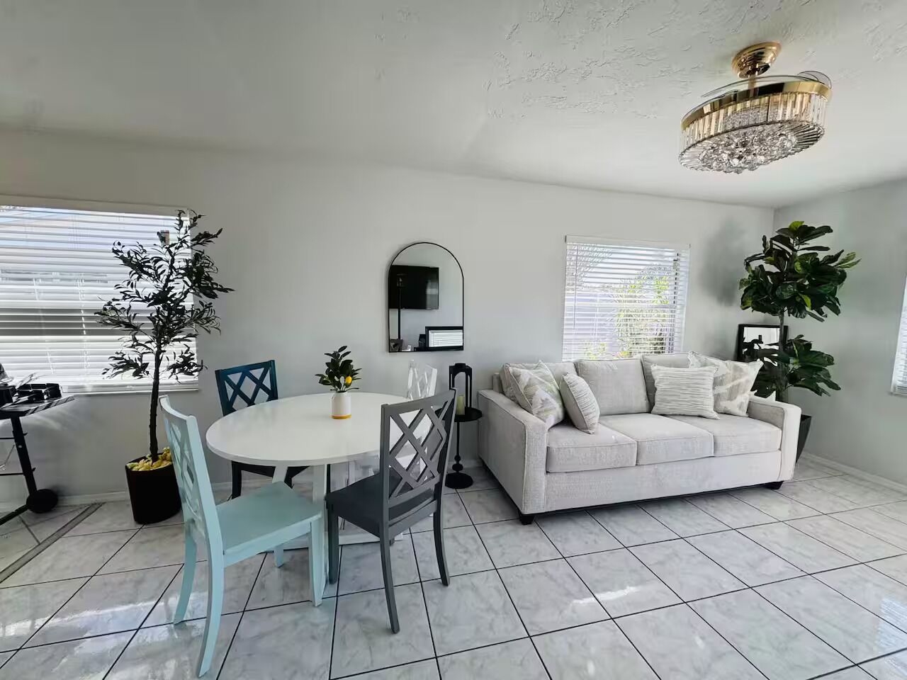 Ein modernes Wohnzimmer mit einem weißen Sofa, einem Esstisch mit Stühlen, einer Kronleuchter und großen Fenstern mit Jalousien.