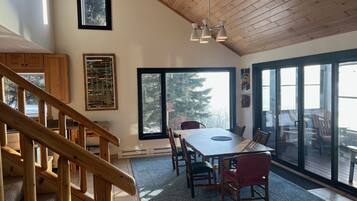 A dining area with a large table, chairs, and a view of the outdoors through large windows.