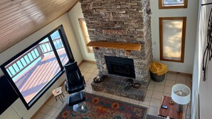 A room with a stone fireplace, a black leather chair, a colorful rug, and a view of a wooden deck through large windows. - Halsnoy Haven New Listing. Iconic property! (Tofte)