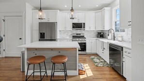 A modern kitchen with white cabinetry, a large island with stools, stainless steel appliances, and pendant lighting. - Modern & Stylish 2/2.5 Home Mainstreet Clayton! (Clayton)