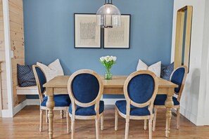Un coin repas avec une table en bois, des chaises à l’assise rembourrée de tissu bleu, un vase avec des fleurs blanches et deux œuvres d’art abstraites encadrées accrochées au mur.