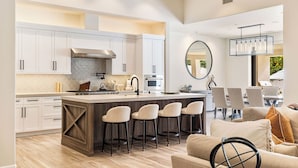 Une cuisine moderne avec des meubles blancs, un îlot central doté de tabourets de bar et un coin repas agrémenté d’un grand miroir rond et de luminaires suspendus.