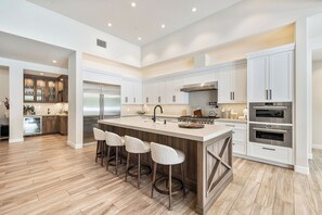 A modern kitchen with a central island, bar stools, stainless steel appliances, and white cabinetry. - Pura Vida - Luxe Estate w/ Pool, Spa & Pickleball! (Rancho Mirage)