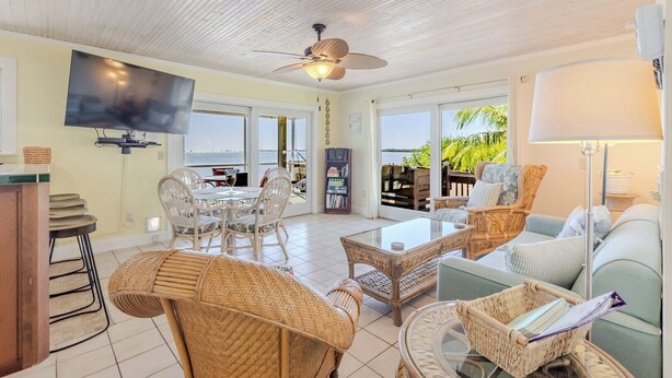 A spacious living area with a television, wicker furniture, a ceiling fan, and large windows offering a view of the outdoors.