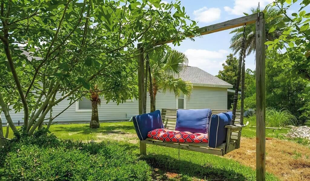 Une balançoire en bois avec un coussin à motifs, dans un jardin luxuriant, avec une maison en arrière-plan.