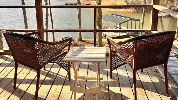 Terraza de madera con sillas de mimbre y una mesa, con vista a un lago y un área boscosa.