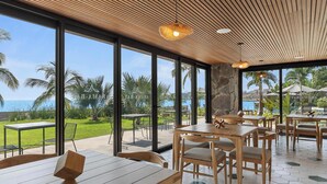 Un vaste espace extérieur aménagé pour les repas, avec des tables et des chaises en bois, de grandes fenêtres offrant une vue sur l’océan et les palmiers, et un plafond en bois agrémenté de luminaires suspendus.