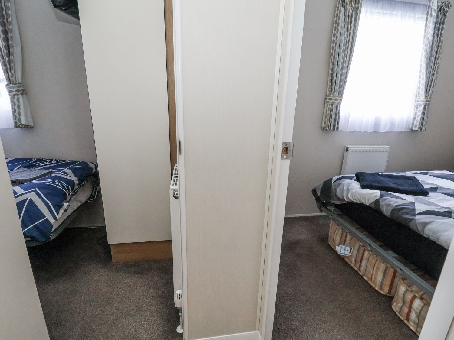 Een kamer met twee bedden, waarvan er een een bedovertrek met dessin heeft en de ander een streepjesbedovertrek. Onder het raam staat een radiator en er is een deur naar een andere kamer.
