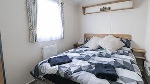 Un dormitorio con una cama, una mesita de noche, una estantería con objetos decorativos y un ventanal con cortinas.