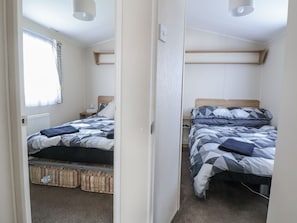 A bedroom with two beds, a window with curtains, a radiator, and a door leading to another room. - Chappie's Caravan 1.0 (Rhyl)
