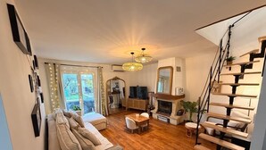 A spacious living room with a sofa, a dining table, and a fireplace. There is a staircase on the right and a large window with curtains. - Family villa in the heart of the Vaucluse (Sorgues)