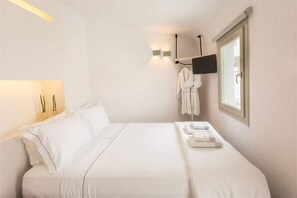 A hotel room with a bed, a flat-screen TV mounted on the wall, a window, and a robe hanging on the wall. - Blessed Blue Sky Jr Suite with heated Jacuzzi and Caldera sea view (Oia)