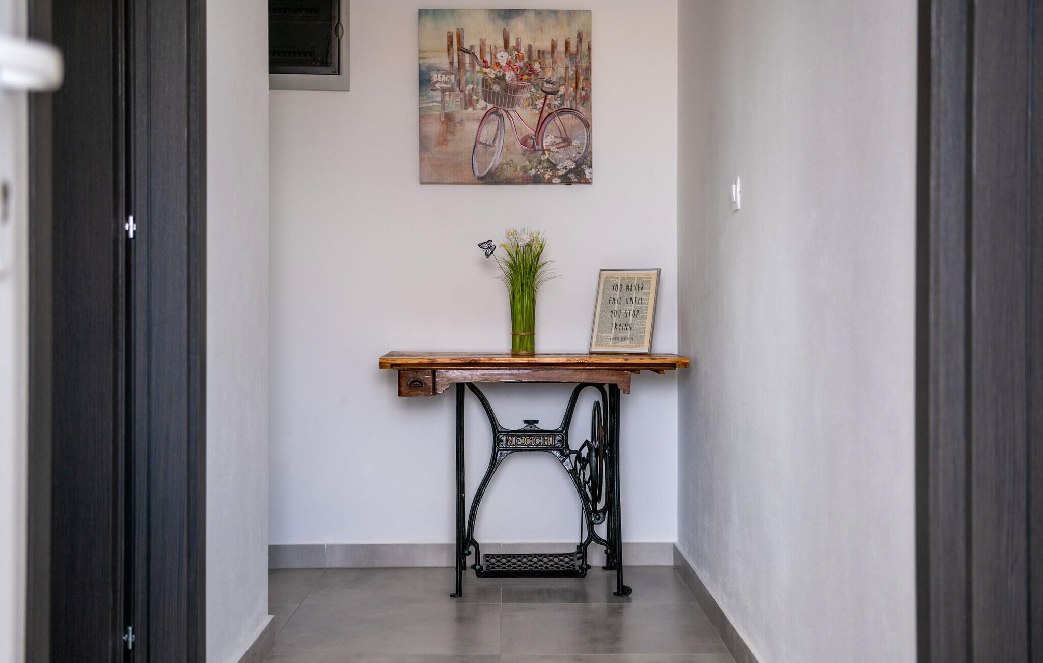 A hallway with a wooden console table, a vase with green plants, a framed inspirational quote, and a vintage sewing machine.