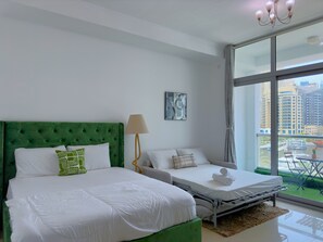 Ein modernes Schlafzimmer mit einem grünen, gepolsterten Kopfteil, zwei Betten, einem Nachttisch, einer Lampe und einem großen Fenster mit Blick auf Gebäude und ein Gewässer.