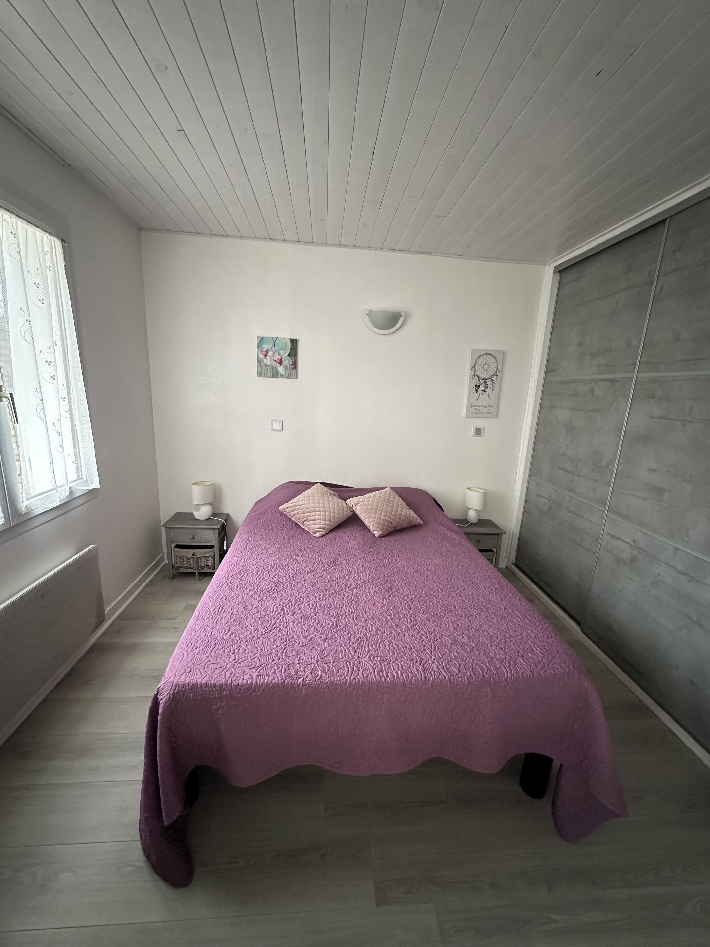 A bedroom with a bed, two nightstands, a window with curtains, a radiator, and a gray wardrobe.