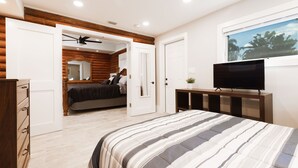 A bedroom with a bed, a TV, a ceiling fan, and a window with a view of trees. - The Cabin – 3BR Log Retreat w/Outdoor Bar & Grill (Jupiter)