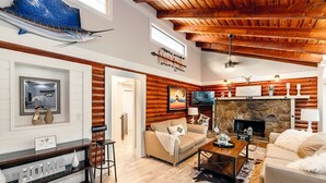 A cozy living room with a stone fireplace, wooden beams, and a television. - The Cabin – 3BR Log Retreat w/Outdoor Bar & Grill (Jupiter)