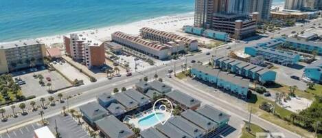 An aerial view of a coastal hotel area with multiple buildings, a parking lot, and a pool.