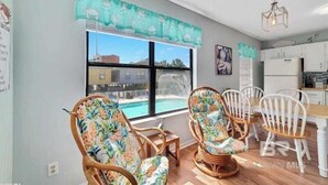 A dining area with wicker chairs and a wooden table, a window with a view of a swimming pool, and a kitchen with white cabinets. - Pet Friendly Beach Access Hangout Nearby Sleeps 6 (Gulf Shores)