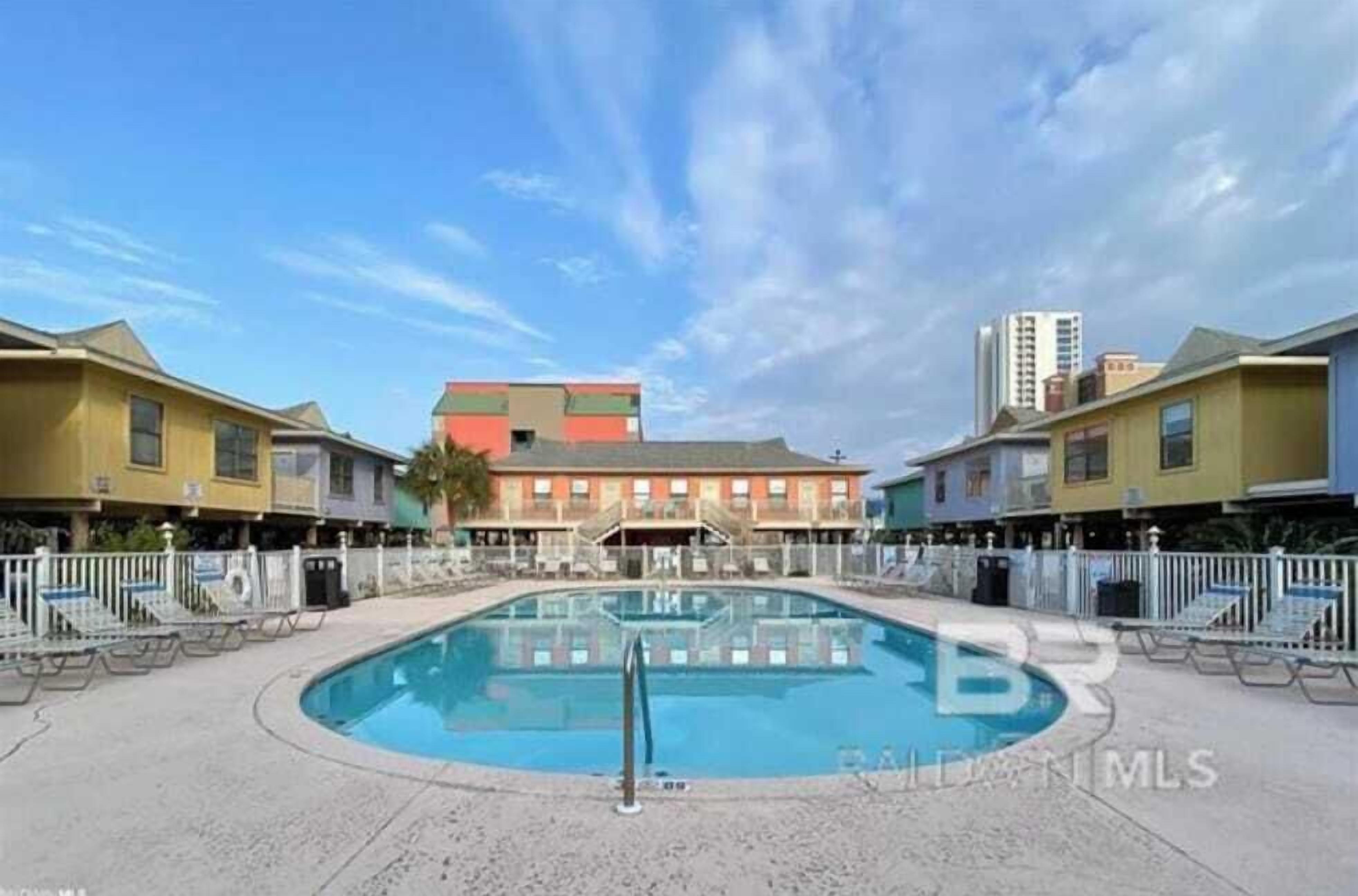 Une piscine entourée d’immeubles à plusieurs étages dotés de balcons, un ciel dégagé et une fontaine d’eau bien visible.