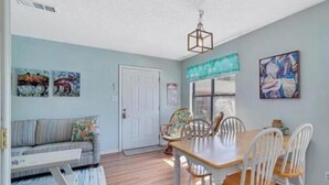A dining area with a wooden table and white chairs, a sofa with cushions, and artwork on the walls. - Pet Friendly Beach Access Hangout Nearby Sleeps 6 (Gulf Shores)
