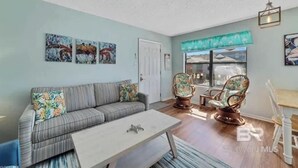 A living room with a sofa, coffee table, and two chairs. There are paintings on the wall and a window with a view. - Pet Friendly Beach Access Hangout Nearby Sleeps 6 (Gulf Shores)