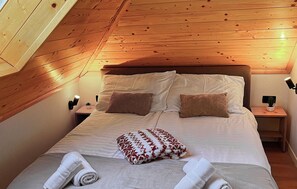 Ein gemütliches Schlafzimmer mit Holzbalkendecke, einem großen Bett mit weißer und brauner Bettwäsche, zwei Nachttischen mit Lampen sowie ein kleiner Topfpflanze auf einem Tisch.