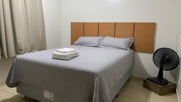 A neatly made bed with a grey quilt, a folded white towel, and a black standing fan. - Hospedaria Estadia Do Vale  (União da Vitória)