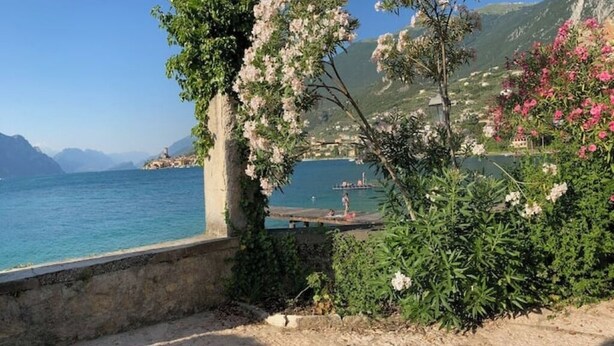 A coastal scene with a stone wall, flowering plants, and a view of the sea and distant mountains. - Sopri House - Relaxation and Lake View, Private Garden and Veranda (Malcesine)