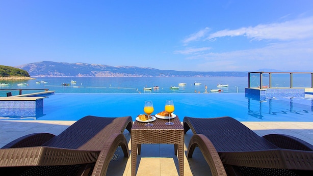 Un espace aménagé au bord de la piscine, avec une table dressée pour le petit-déjeuner, offrant une vue sur la mer, avec des bateaux et des collines à l’horizon.